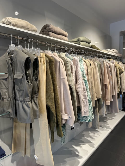 A rack of neutral-toned clothing displayed in a store, including sweaters, jackets, and folded knitwear in shades of beige, cream, gray, and pastel green.